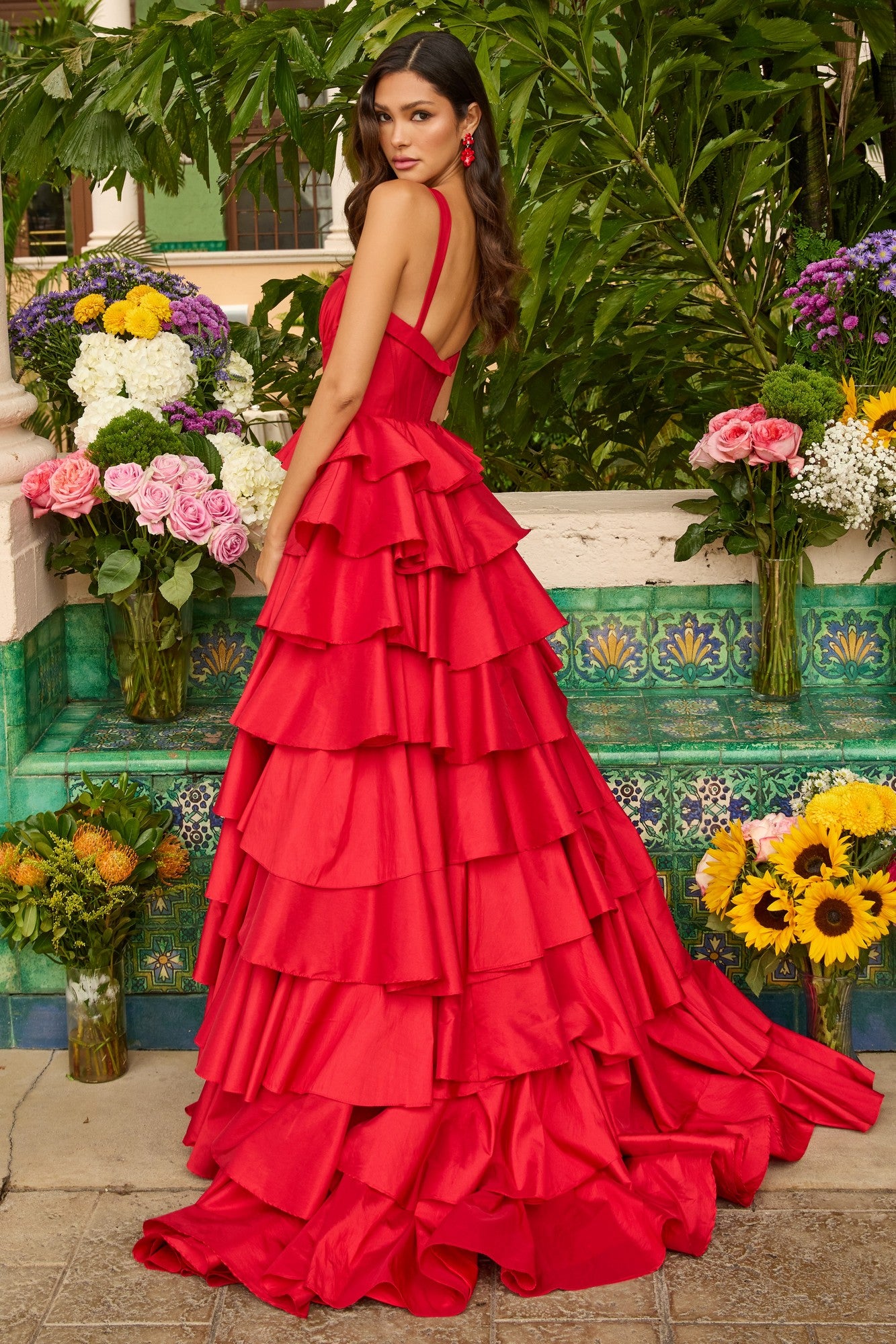 Outdoors among flowers, a woman wears the Ava Presley 42234 long prom dress—a bright red gown with a tiered ruffle skirt, open back, corset bodice, and matching red earrings.